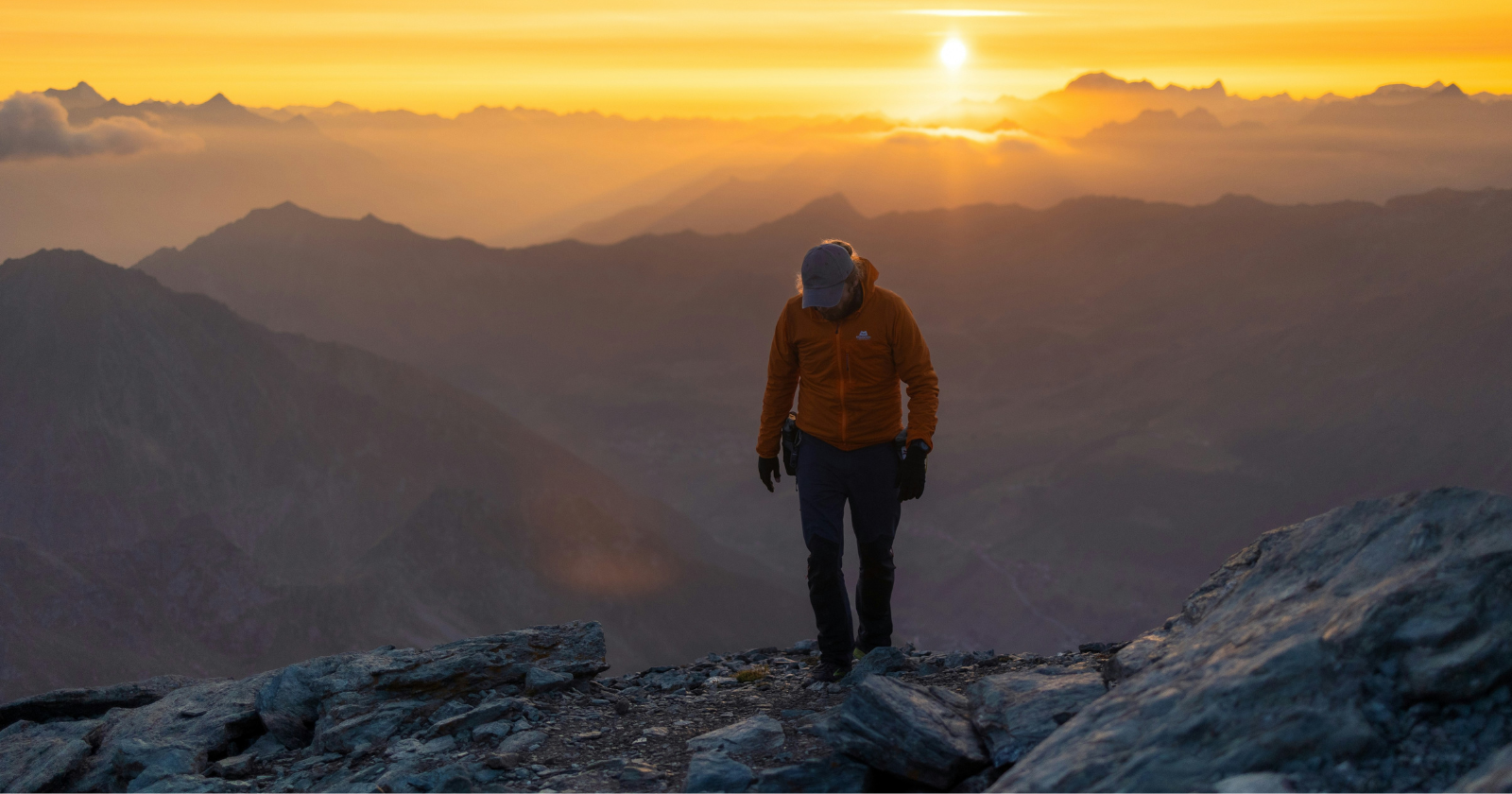 A hiker in an orange jacket and cap walks up a rocky summit at sunrise, with layers of mountains and a low sun behind