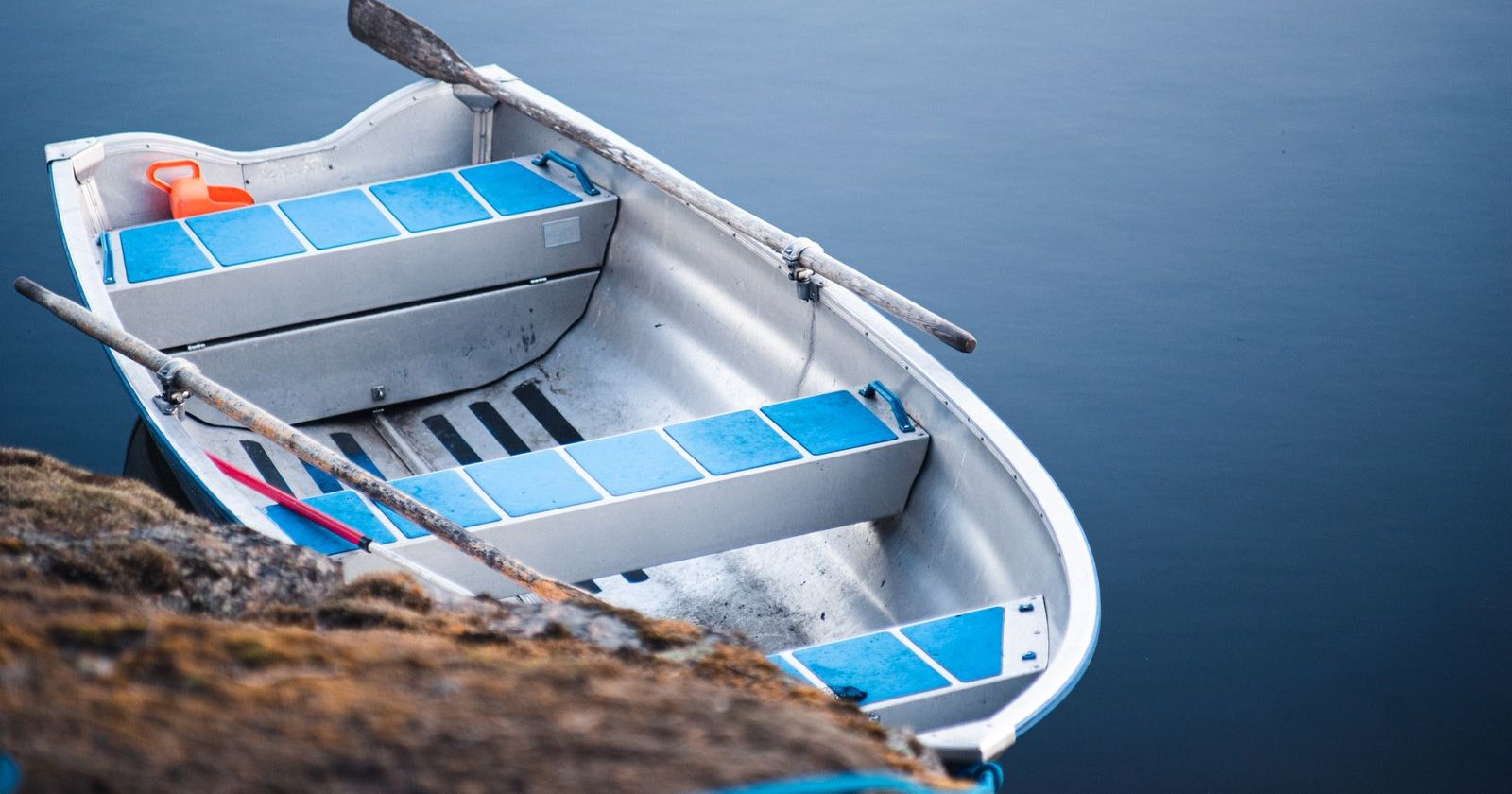 An empty rowboat with oars resting on calm blue water beside a rocky shore