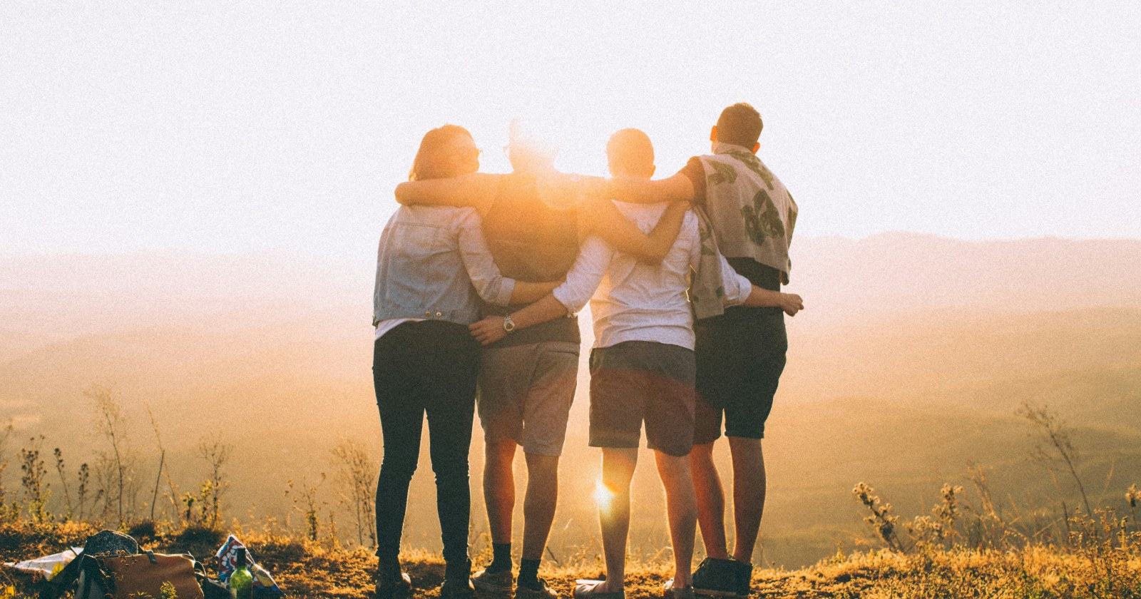 Four friends seen from behind, arms around each other's shoulders, looking out over hills at sunset