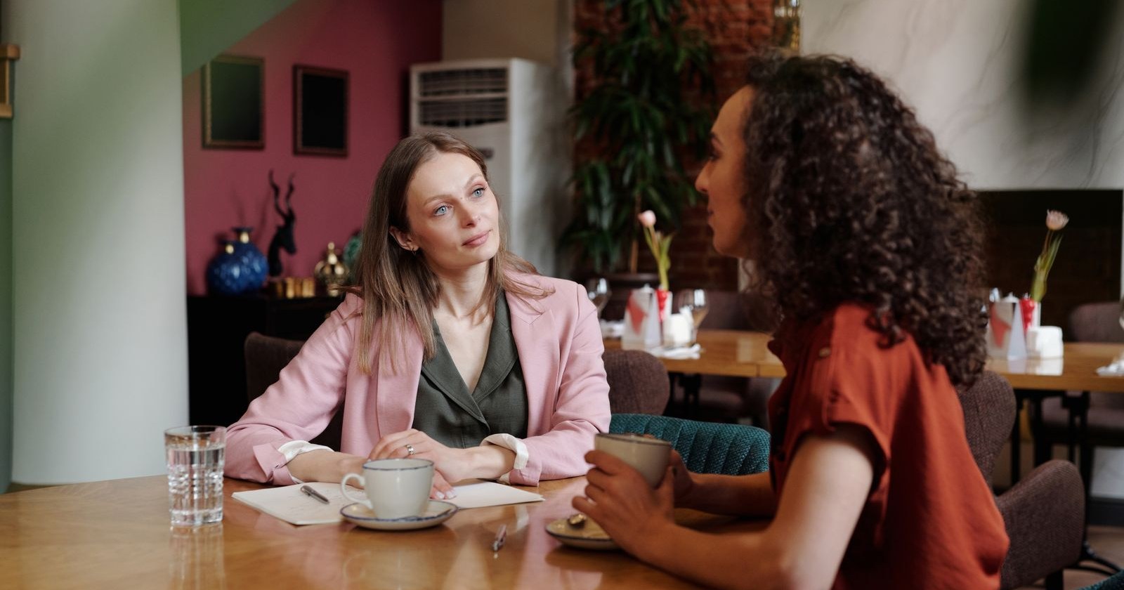 Two women talking over coffee at a cafe table, one listening with a thoughtful, slightly guarded expression