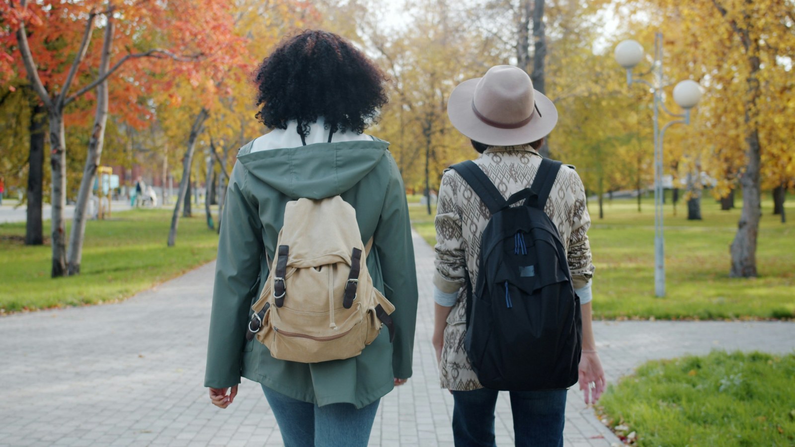 Two friends with backpacks walking side by side along a park path in autumn