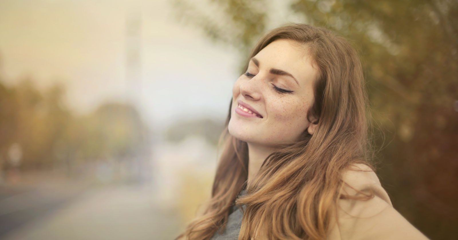 A woman smiling with her eyes closed outdoors, looking relaxed and at ease