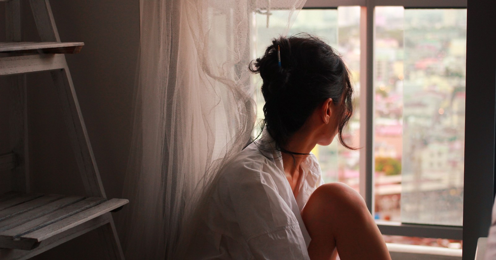 A woman in a white shirt sits by a tall window, looking out over a city