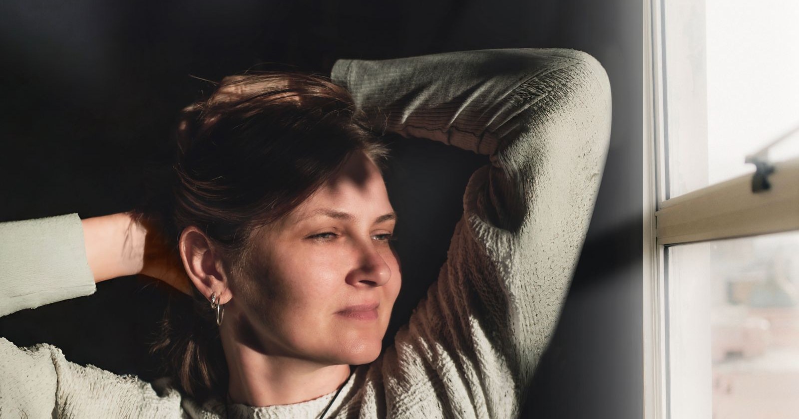 A woman relaxing with her hands behind her head, looking out a sunlit window
